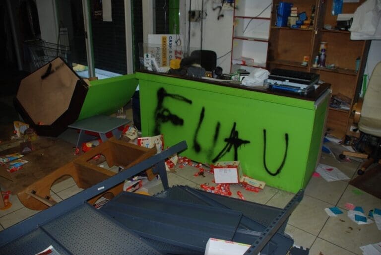A vandalized store interior with a neon green counter spray-painted with black graffiti. While the floor is covered in discarded products and knocked-over shelving.