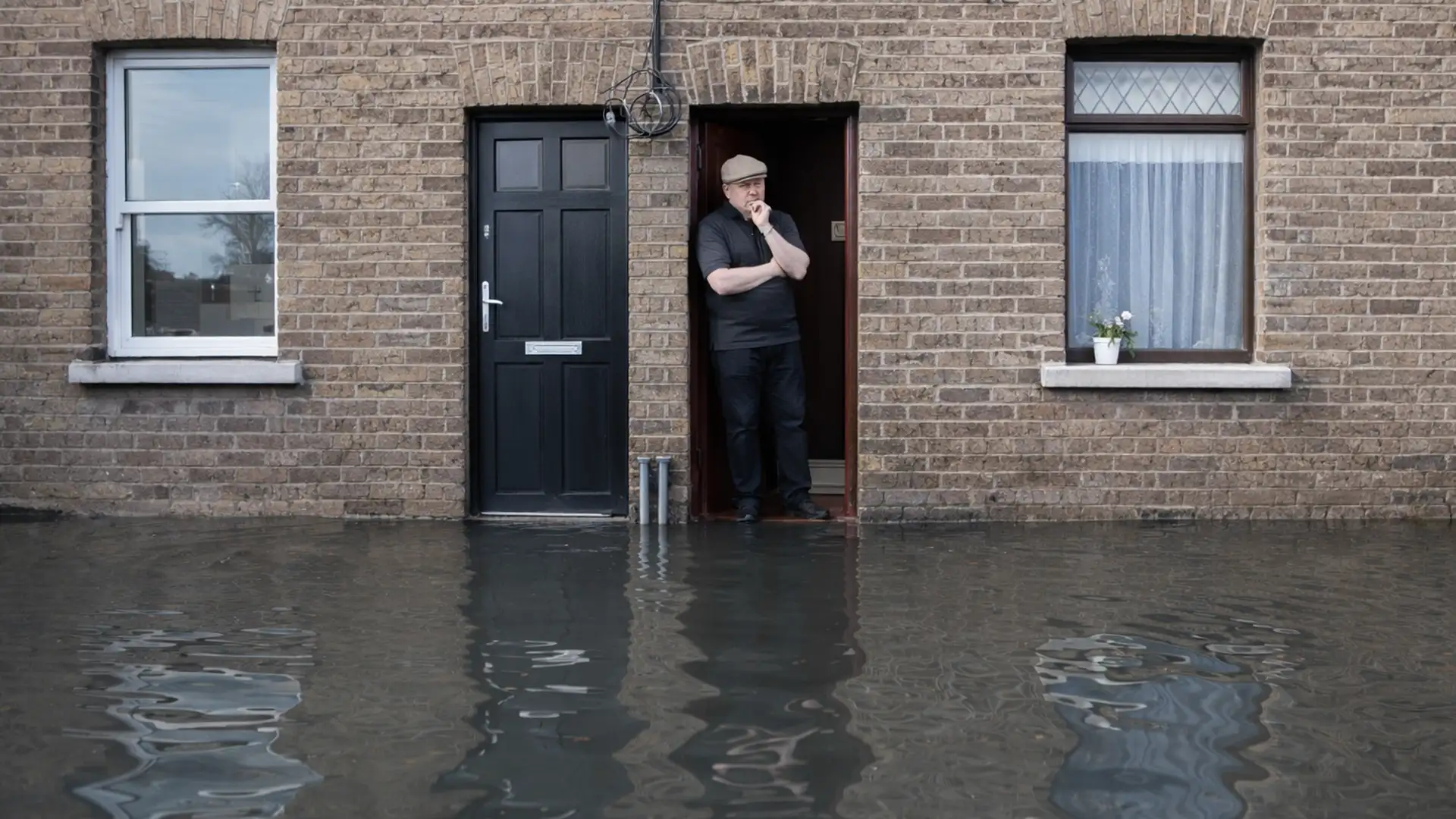 The man is concerned, standing in the open doorway of a brown brick building. Deep, dark floodwater covers the entire foreground and street, reflecting the building's windows, doors and the sky.